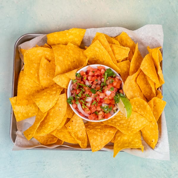 A metal tray filled with crispy tortilla chips and a white bowl of fresh salsa with a lime wedge.