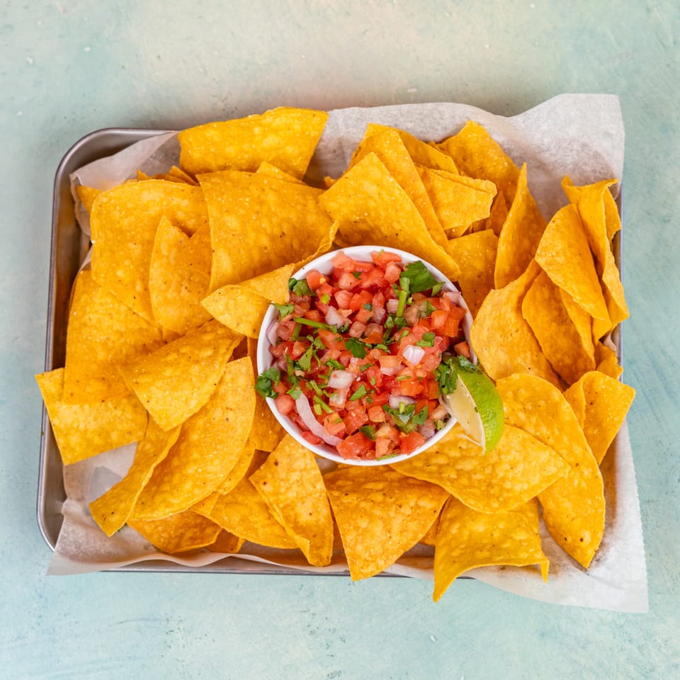 A metal tray filled with crispy tortilla chips and a white bowl of fresh salsa with a lime wedge.