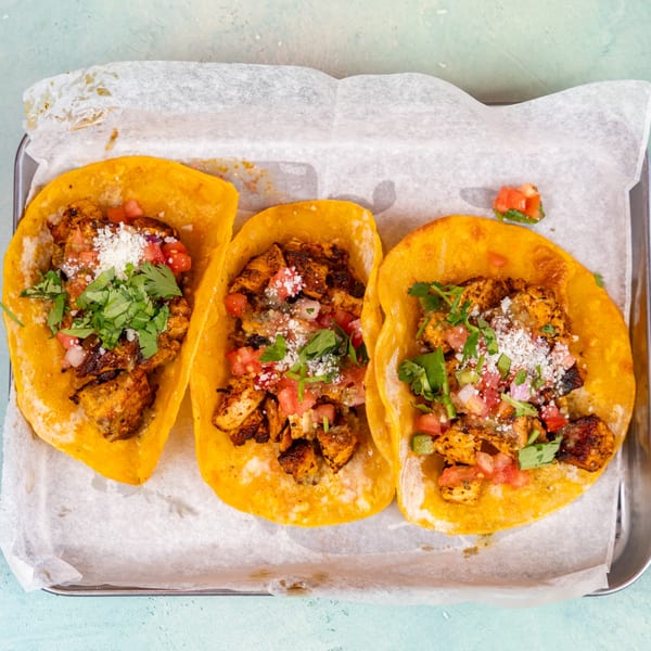 Three chicken tacos with pico de gallo, cilantro, and cotija cheese on a metal tray.