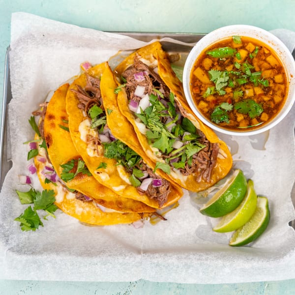 Birria tacos with cilantro, onions, a side of consommé, and lime wedges on a tray.