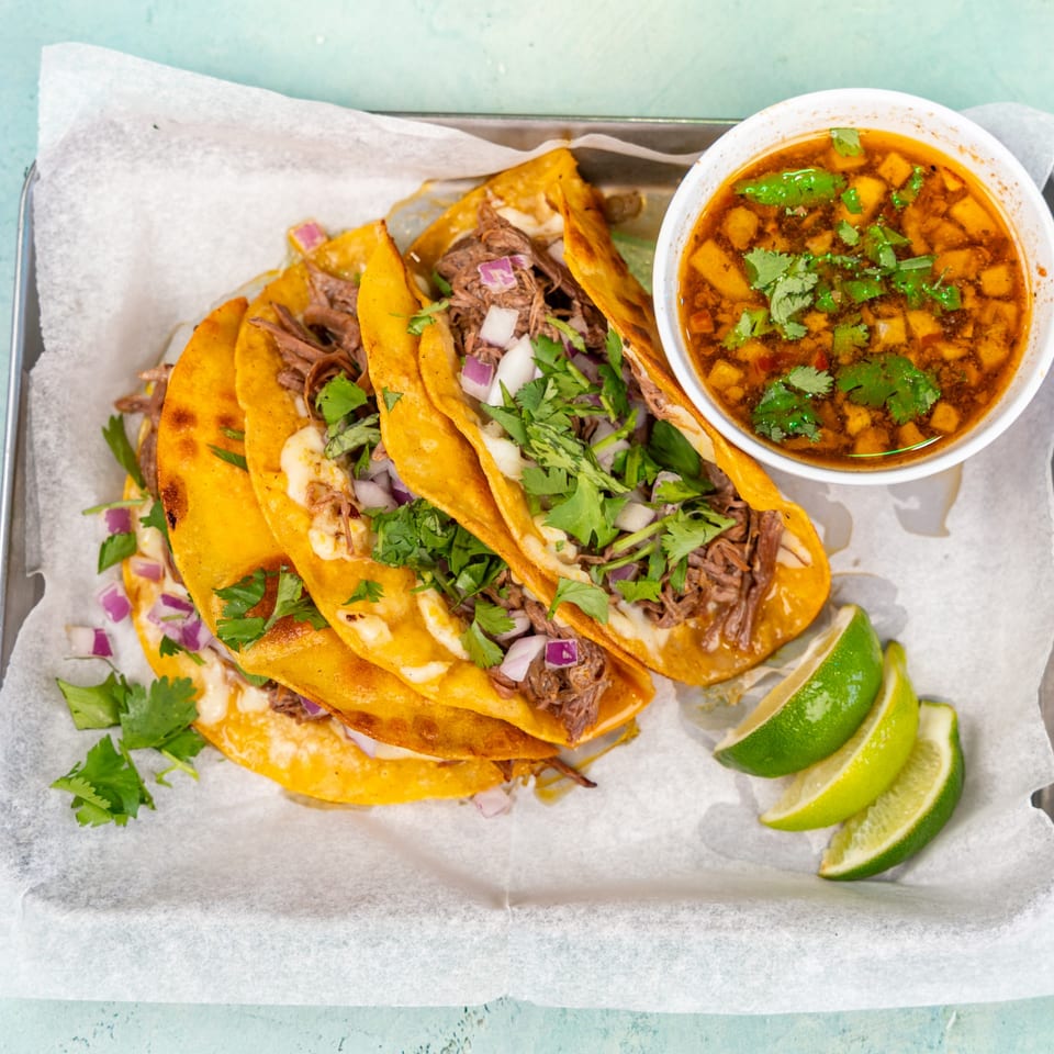 Birria tacos with cilantro, onions, a side of consommé, and lime wedges on a tray.