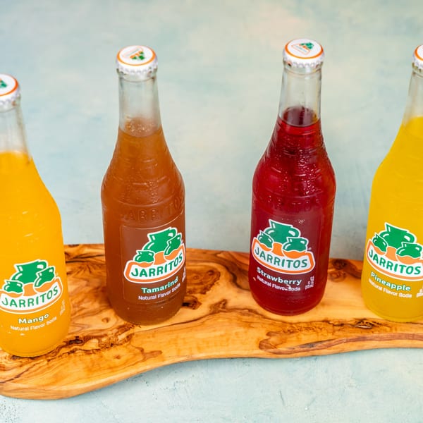 Four Jarritos soda bottles in mango, tamarind, strawberry, and pineapple flavors on a wooden board.