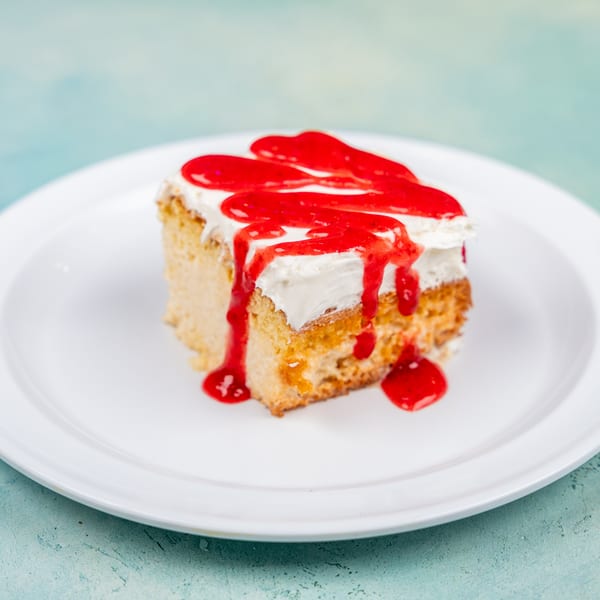 A slice of tres leches cake with white frosting and red strawberry syrup on a white plate.
