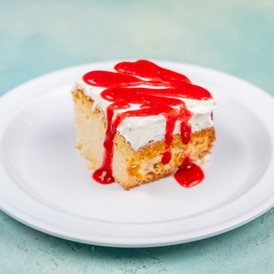 A slice of tres leches cake with white frosting and red strawberry syrup on a white plate.