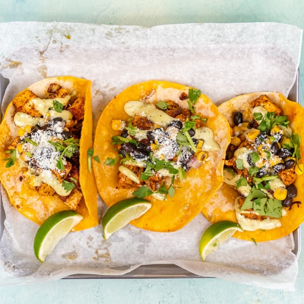 Three chicken tacos with black beans, corn, cilantro, and lime wedges on a baking sheet.