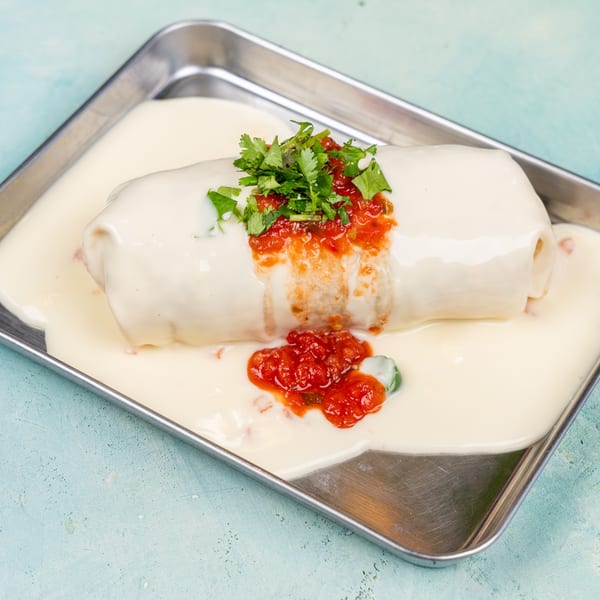 A large burrito covered in white cheese sauce, topped with red salsa and fresh cilantro on a metal tray.