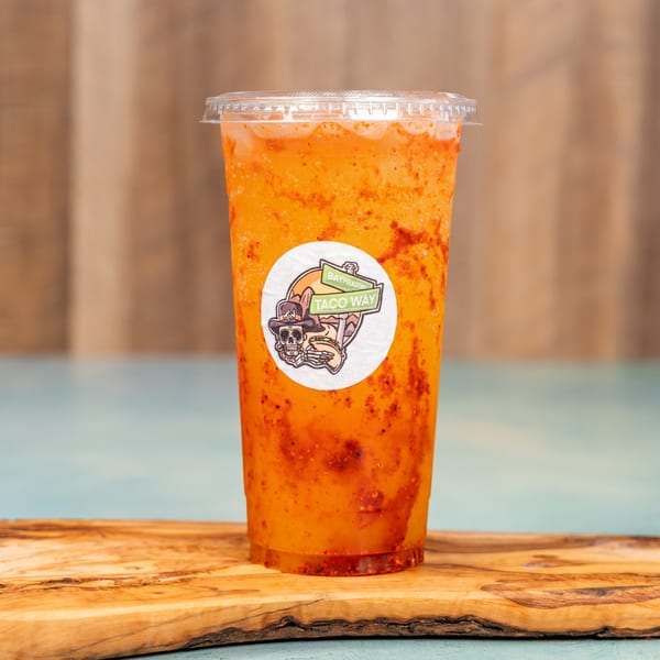 A vibrant orange frozen drink with red spices, featuring a "Taco Way" logo, on a wooden board.