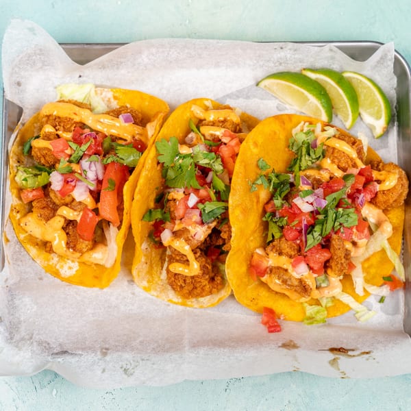 Three crispy shrimp tacos with sauce, pico de gallo, and cilantro, served with lime wedges.