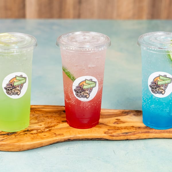 Three colorful Taco Way sodas in plastic cups with brand stickers on a wooden board.