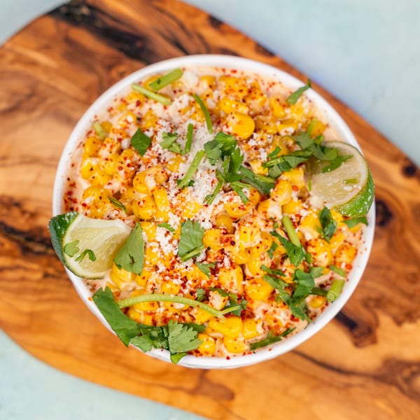 Elote, Mexican street corn, in a white bowl with cilantro, chili powder, and lime wedges.