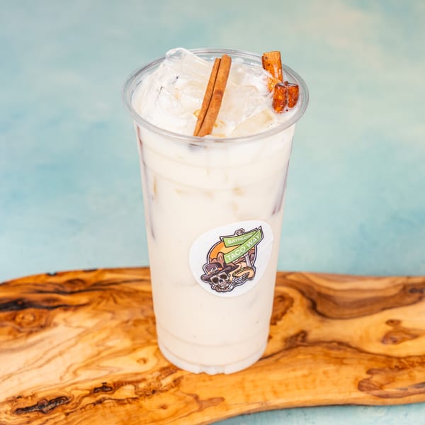Horchata drink in a clear plastic cup with ice and cinnamon sticks, on a wooden board.