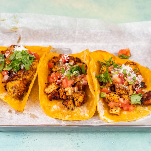 Three chicken tacos with pico de gallo, cilantro, and cheese on a metal tray.