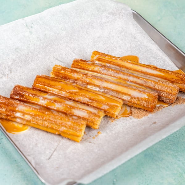 Golden churros drizzled with white icing and caramel sauce, sprinkled with cinnamon sugar on parchment paper.