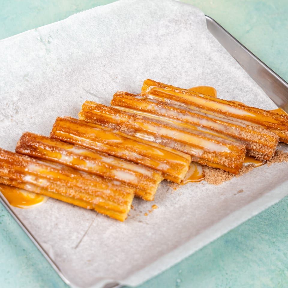 Golden churros drizzled with white icing and caramel sauce, sprinkled with cinnamon sugar on parchment paper.
